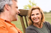 A man and woman are sitting outdoors on wooden chairs, smiling at each other. The woman has shoulder-length hair and is wearing a gray top. The man has short hair and is wearing an orange shirt. They appear to be in a grassy area with trees in the background.