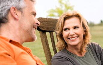 A man and woman are sitting outdoors on wooden chairs, smiling at each other. The woman has shoulder-length hair and is wearing a gray top. The man has short hair and is wearing an orange shirt. They appear to be in a grassy area with trees in the background.