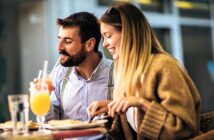 A man and a woman are sitting at an outdoor cafe table. The man is sipping a drink with a straw, while the woman is smiling, holding a fork and knife. They appear to be enjoying a meal together in a relaxed setting.