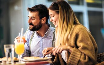 A man and a woman are sitting at an outdoor cafe table. The man is sipping a drink with a straw, while the woman is smiling, holding a fork and knife. They appear to be enjoying a meal together in a relaxed setting.