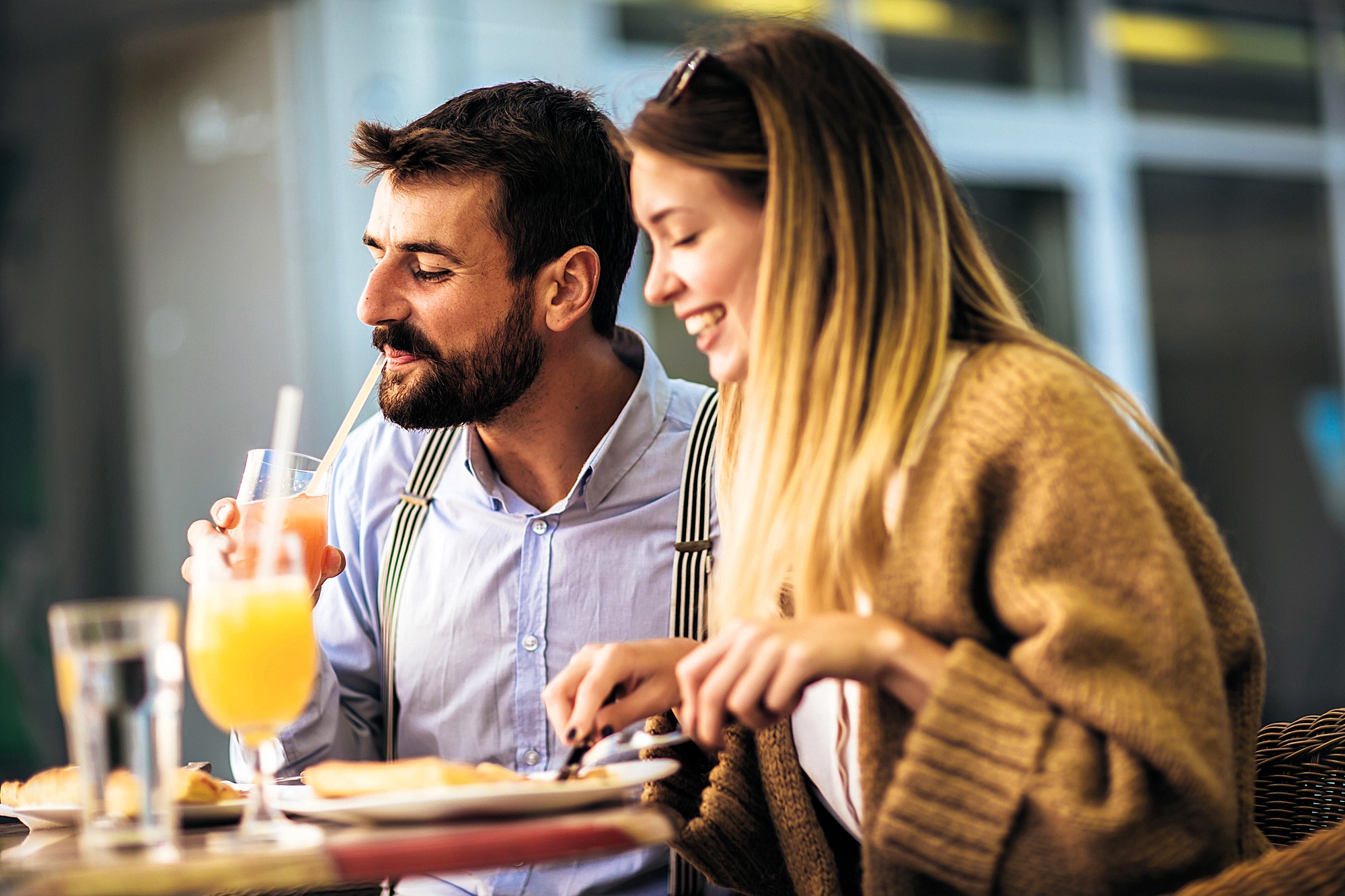 A man and a woman are sitting at an outdoor cafe table. The man is sipping a drink with a straw, while the woman is smiling, holding a fork and knife. They appear to be enjoying a meal together in a relaxed setting.