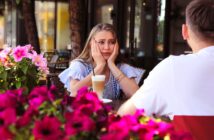 A woman looks upset with her hands on her cheeks while sitting at an outdoor café table across from a man. A glass of latte is in front of her. Pink flowers are in the foreground, and the café setting is lush and vibrant.