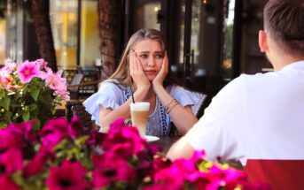 A woman looks upset with her hands on her cheeks while sitting at an outdoor café table across from a man. A glass of latte is in front of her. Pink flowers are in the foreground, and the café setting is lush and vibrant.