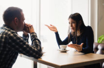 A woman and a man are sitting across from each other at a café table. The woman gestures with both hands, appearing frustrated. The man listens with his hands clasped near his face. Coffee cups are on the table. Natural light fills the space.