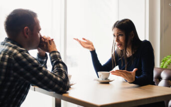 A woman and a man are sitting across from each other at a café table. The woman gestures with both hands, appearing frustrated. The man listens with his hands clasped near his face. Coffee cups are on the table. Natural light fills the space.