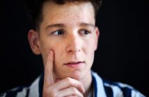 A person with freckles and short curly hair is looking to the side thoughtfully, with their finger resting on their cheek. They are wearing a striped shirt, and the background is dark.