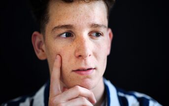 A person with freckles and short curly hair is looking to the side thoughtfully, with their finger resting on their cheek. They are wearing a striped shirt, and the background is dark.