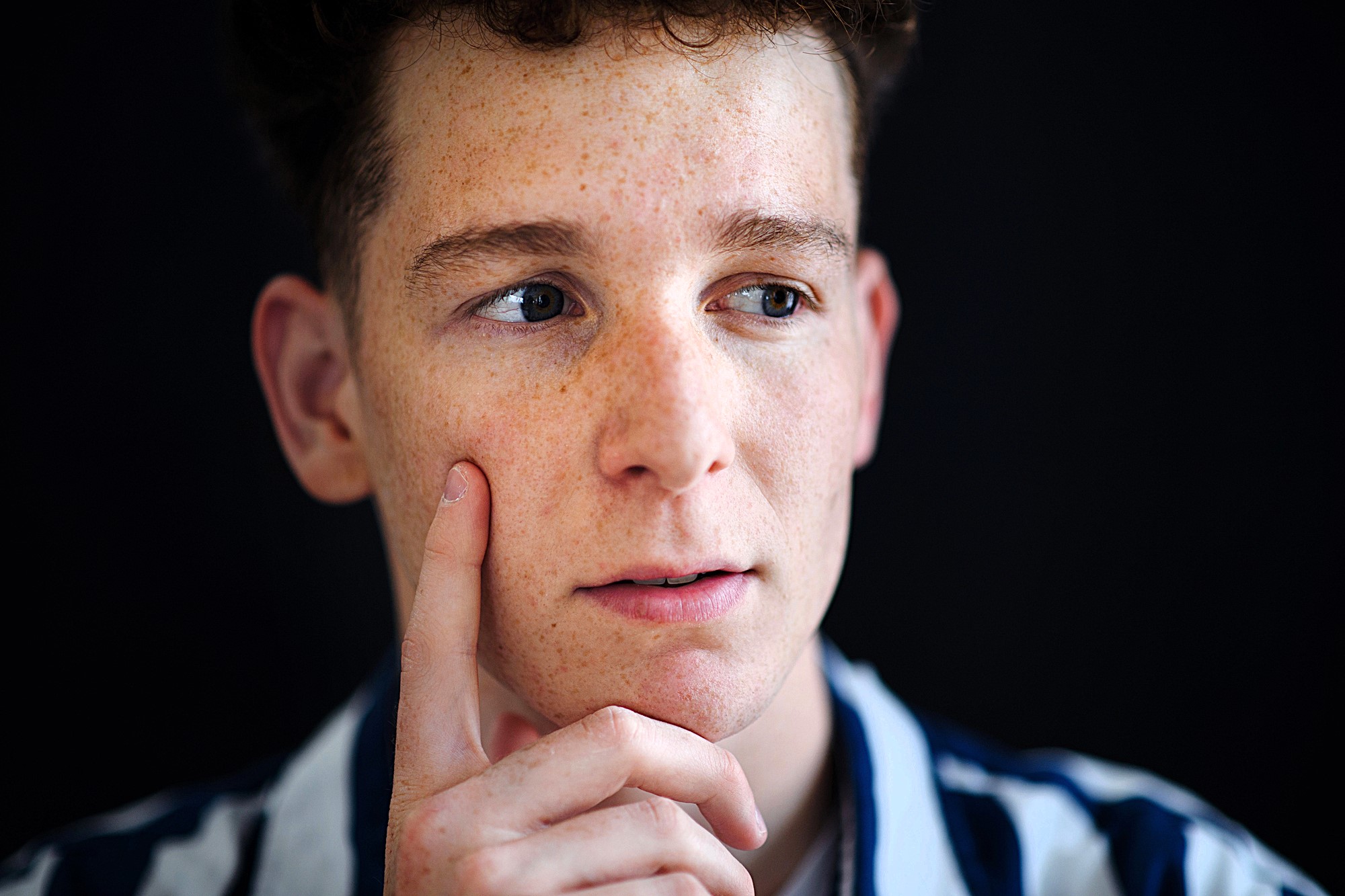 A person with freckles and short curly hair is looking to the side thoughtfully, with their finger resting on their cheek. They are wearing a striped shirt, and the background is dark.