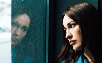 A woman with long dark hair gazes thoughtfully out a window, with her reflection visible on the glass. She appears deep in thought, framed by the window and soft natural light.