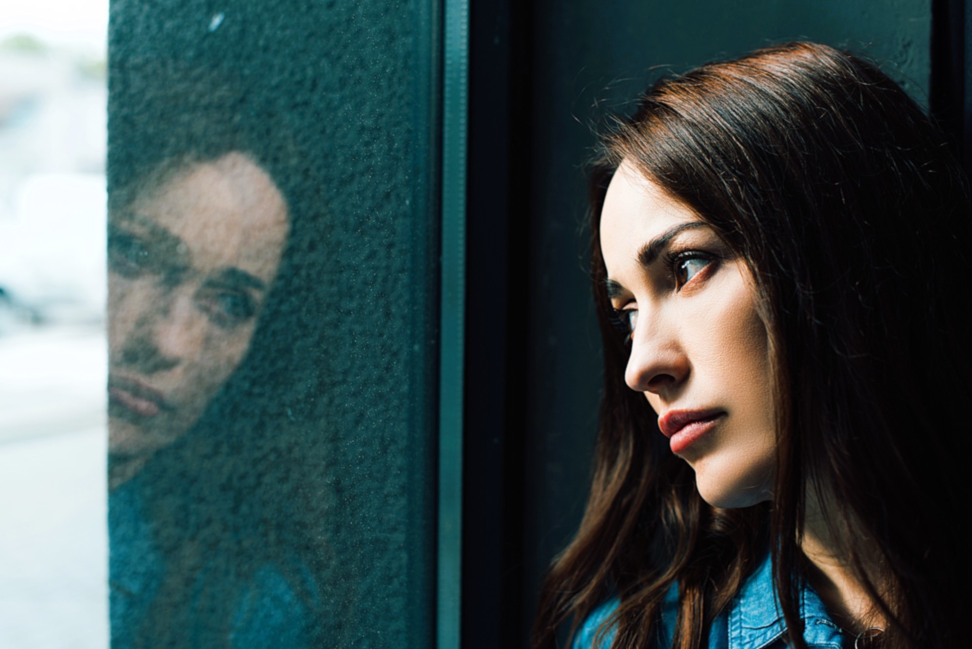 A woman with long dark hair gazes thoughtfully out a window, with her reflection visible on the glass. She appears deep in thought, framed by the window and soft natural light.