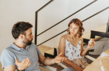 A man and a woman are sitting on stairs at home, engaged in a conversation. The man gestures with his hands while speaking, and the woman holds a sandwich, listening attentively. They're in a casual, modern setting with a railing in the background.