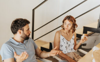 A man and a woman are sitting on stairs at home, engaged in a conversation. The man gestures with his hands while speaking, and the woman holds a sandwich, listening attentively. They're in a casual, modern setting with a railing in the background.