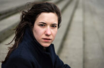 A woman with long brown hair looks intently at the camera. She is wearing a dark coat. The background is blurred, showing steps leading downward.