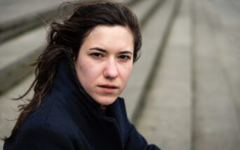 A woman with long brown hair looks intently at the camera. She is wearing a dark coat. The background is blurred, showing steps leading downward.