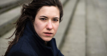 A woman with long brown hair looks intently at the camera. She is wearing a dark coat. The background is blurred, showing steps leading downward.