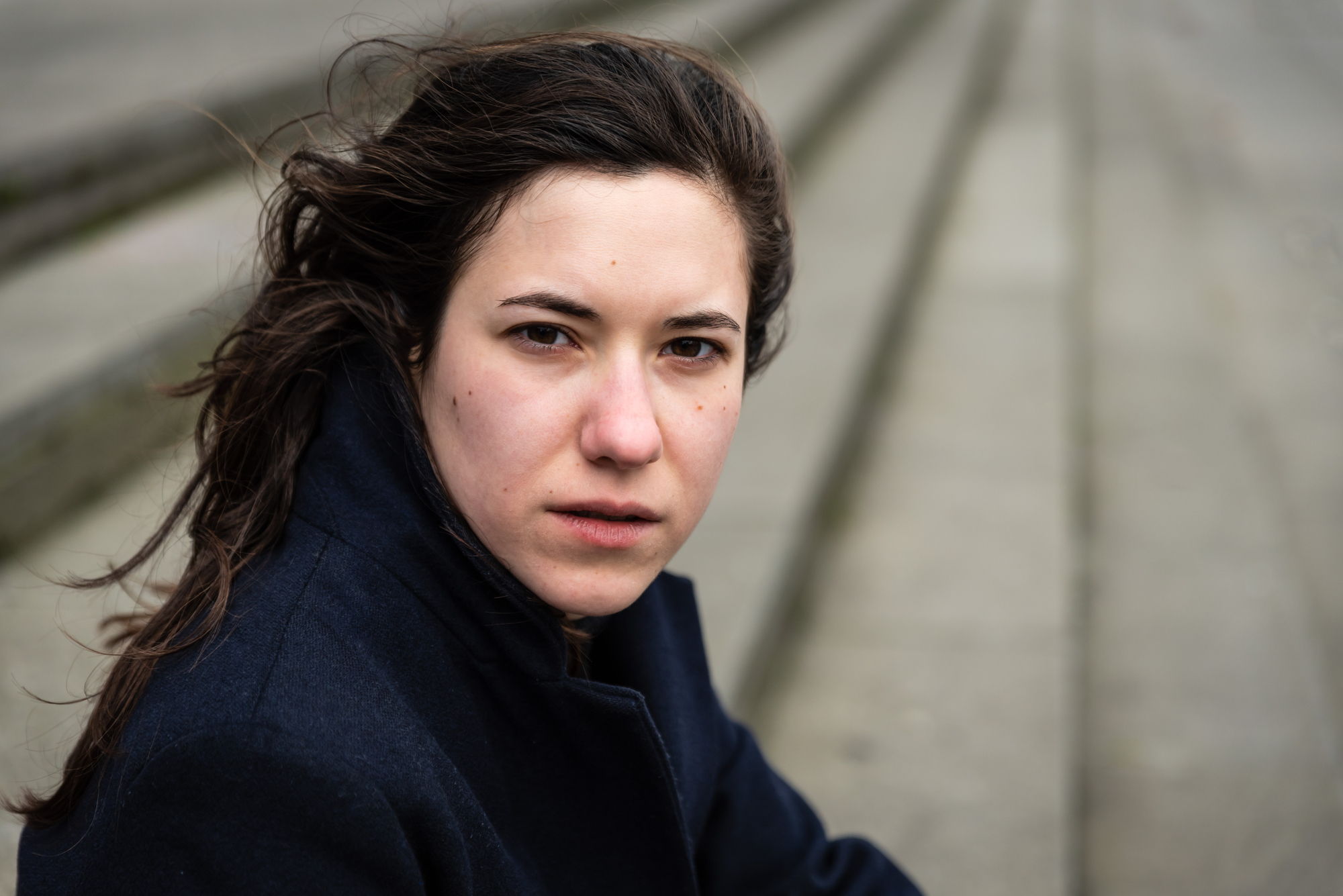 A woman with long brown hair looks intently at the camera. She is wearing a dark coat. The background is blurred, showing steps leading downward.