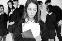 A girl stands alone, looking down and holding books. In the background, a group of students in uniforms are whispering and pointing, creating a sense of exclusion. The image is in black and white.