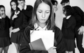 A girl stands alone, looking down and holding books. In the background, a group of students in uniforms are whispering and pointing, creating a sense of exclusion. The image is in black and white.