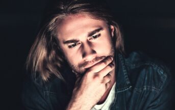 A person with long hair and a beard is deep in thought, resting their chin on their hand. They are wearing a denim jacket and a white shirt, with a dark background emphasizing their contemplative expression.