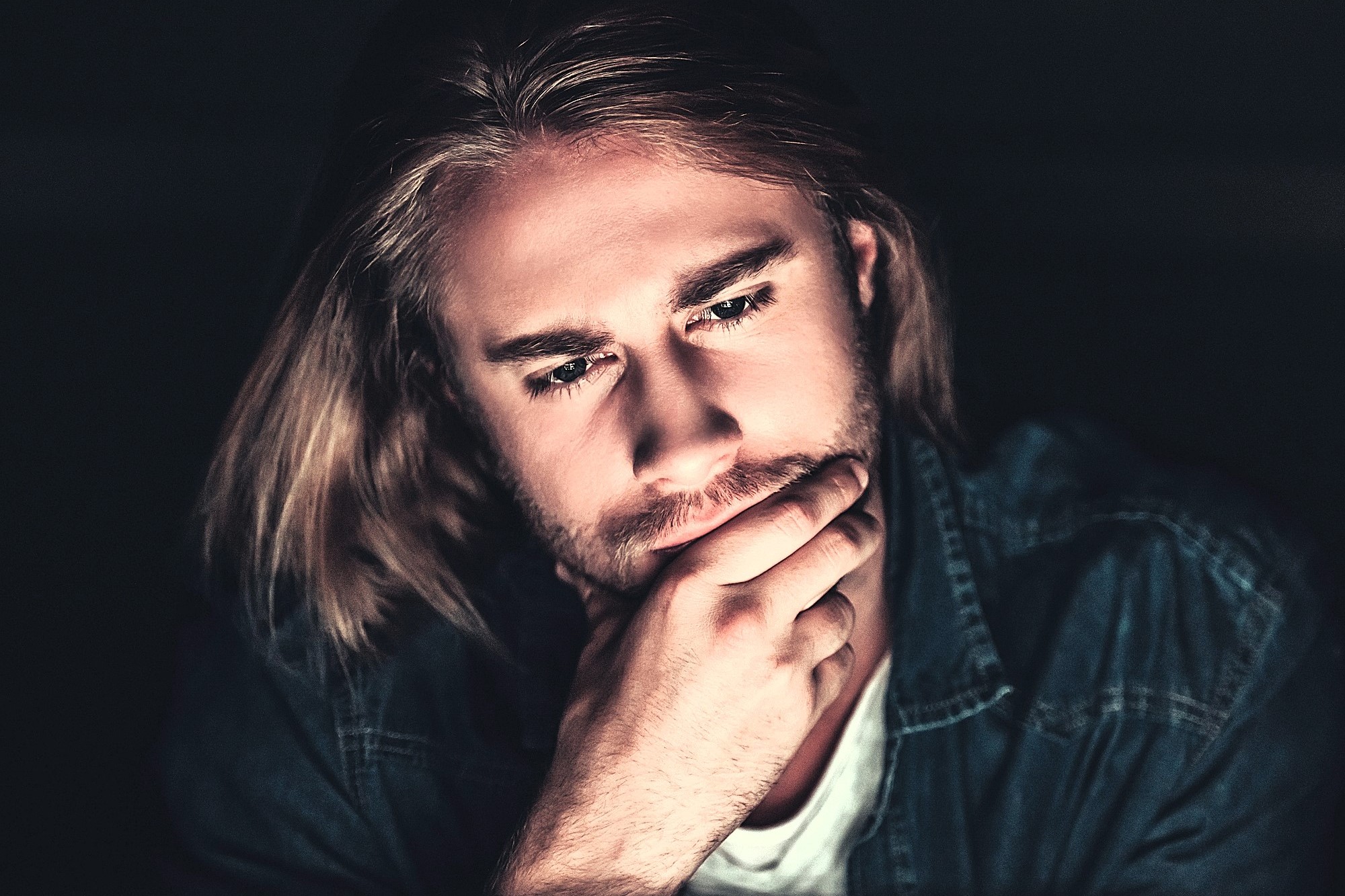 A person with long hair and a beard is deep in thought, resting their chin on their hand. They are wearing a denim jacket and a white shirt, with a dark background emphasizing their contemplative expression.