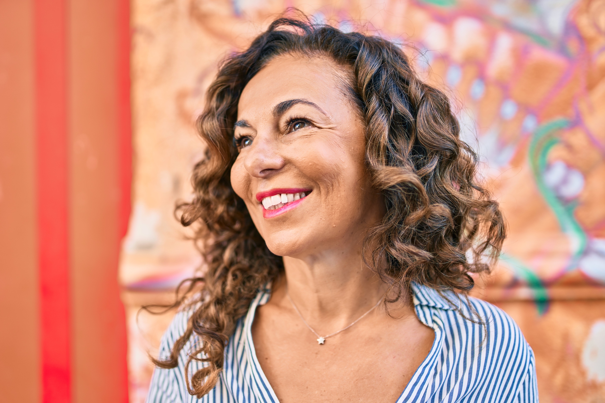 A woman with curly hair smiles while looking off-camera. She is wearing a striped shirt and a delicate necklace. The background features a colorful, abstract mural.