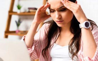 A woman in a striped shirt sits in front of a laptop, massaging her temples with a pained expression, suggesting stress or a headache. Books and plants are blurred in the background on a wooden shelf.