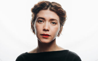 Portrait of a person with short, wavy hair against a white background. They are wearing a black top and have a neutral expression, with light makeup and lipstick.