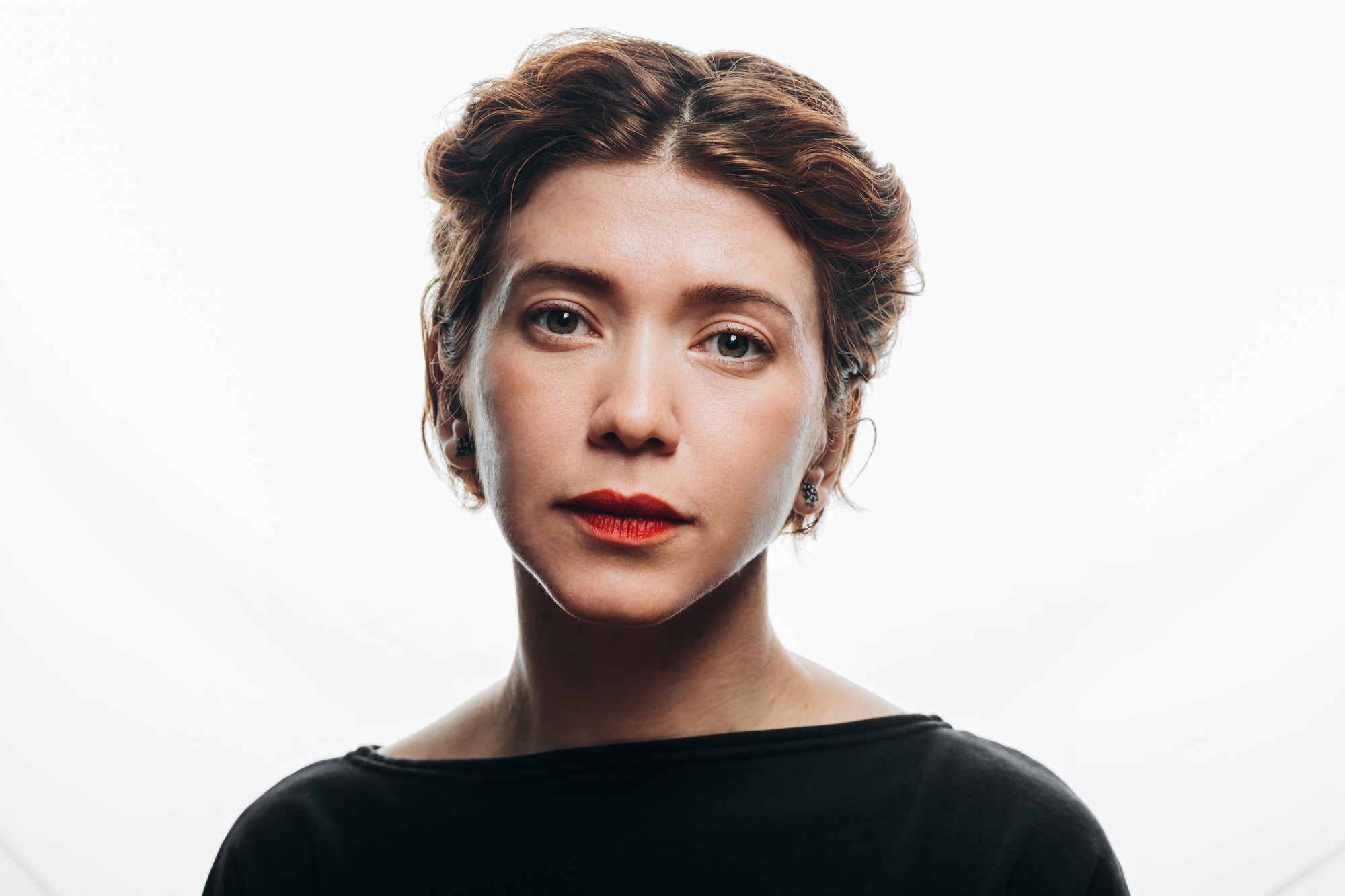 Portrait of a person with short, wavy hair against a white background. They are wearing a black top and have a neutral expression, with light makeup and lipstick.