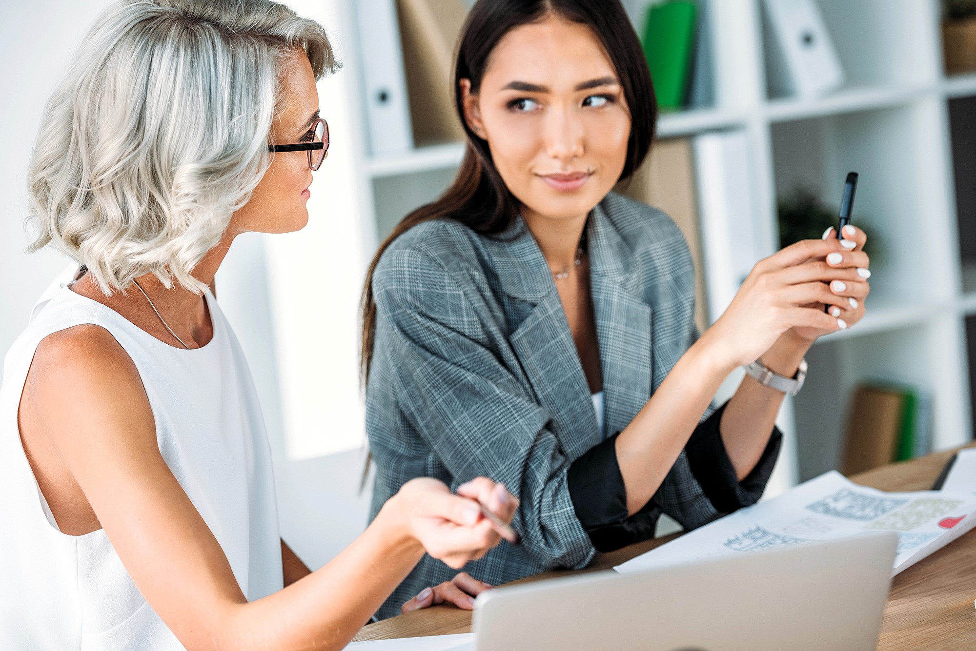 Two women are sitting at a desk in an office. One, with gray hair and glasses, is speaking, while the other, with long dark hair, listens intently, holding a pen. Papers and a laptop are on the desk in front of them. Shelves are in the background.