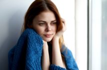 A woman with long brown hair looks thoughtfully out of a window, wrapped in a blue knitted blanket. The natural light highlights her contemplative expression.