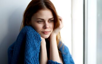 A woman with long brown hair looks thoughtfully out of a window, wrapped in a blue knitted blanket. The natural light highlights her contemplative expression.