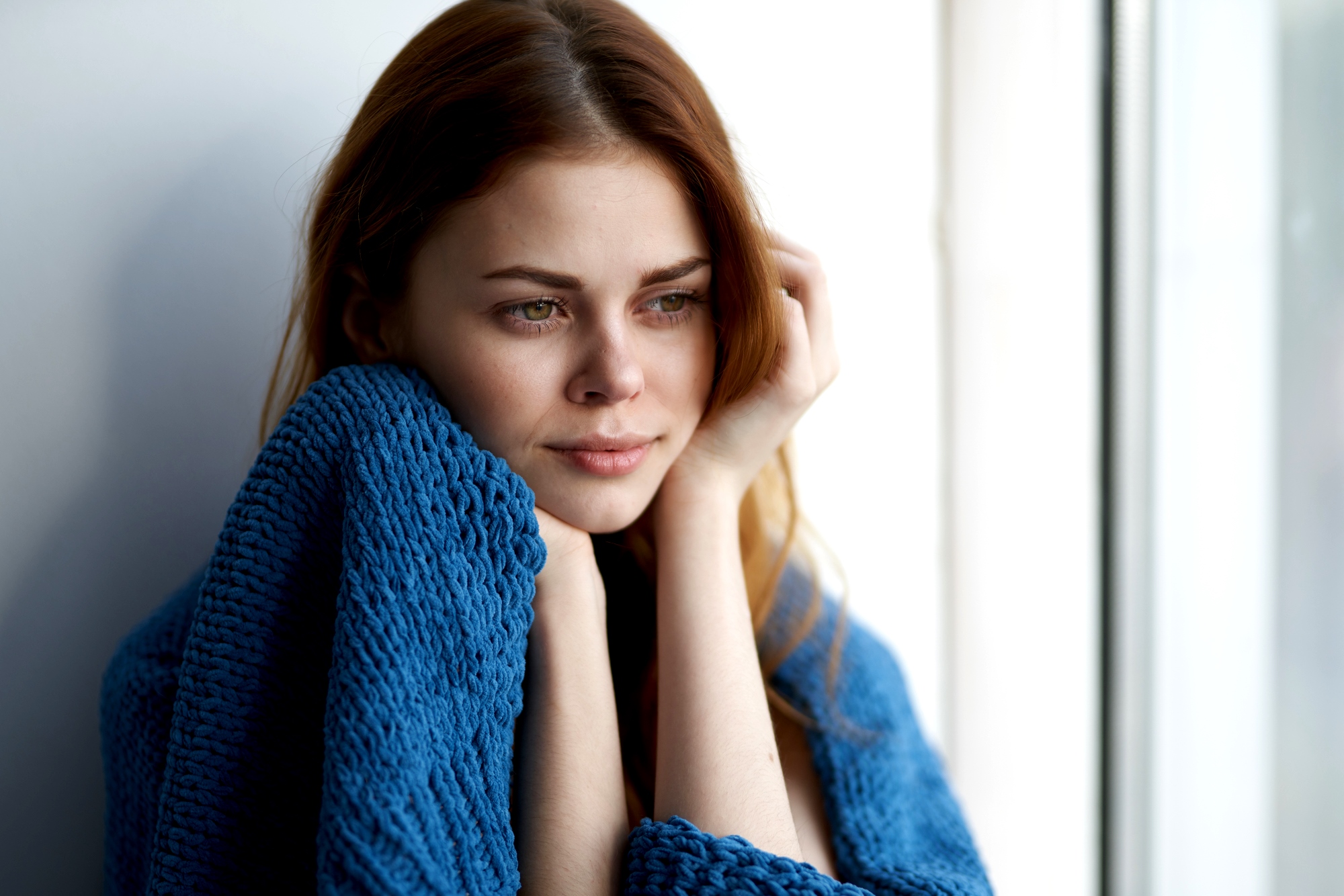 A woman with long brown hair looks thoughtfully out of a window, wrapped in a blue knitted blanket. The natural light highlights her contemplative expression.