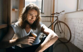 A person with long hair sits on the floor, holding a coffee cup and looking down thoughtfully. A bicycle and a window are in the background, with light streaming into the room.