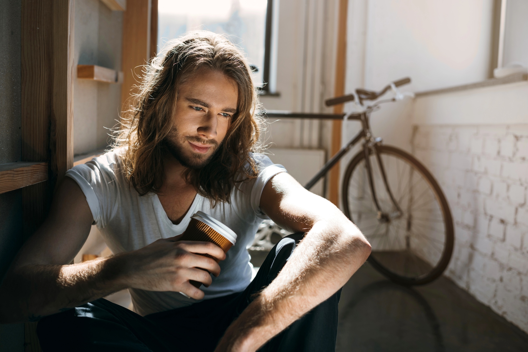 A person with long hair sits on the floor, holding a coffee cup and looking down thoughtfully. A bicycle and a window are in the background, with light streaming into the room.