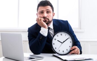 A man in a suit sits at a desk, resting his head on one hand and holding a wall clock with the other. A laptop and clipboard are in front of him. The expression on his face appears bored or impatient.