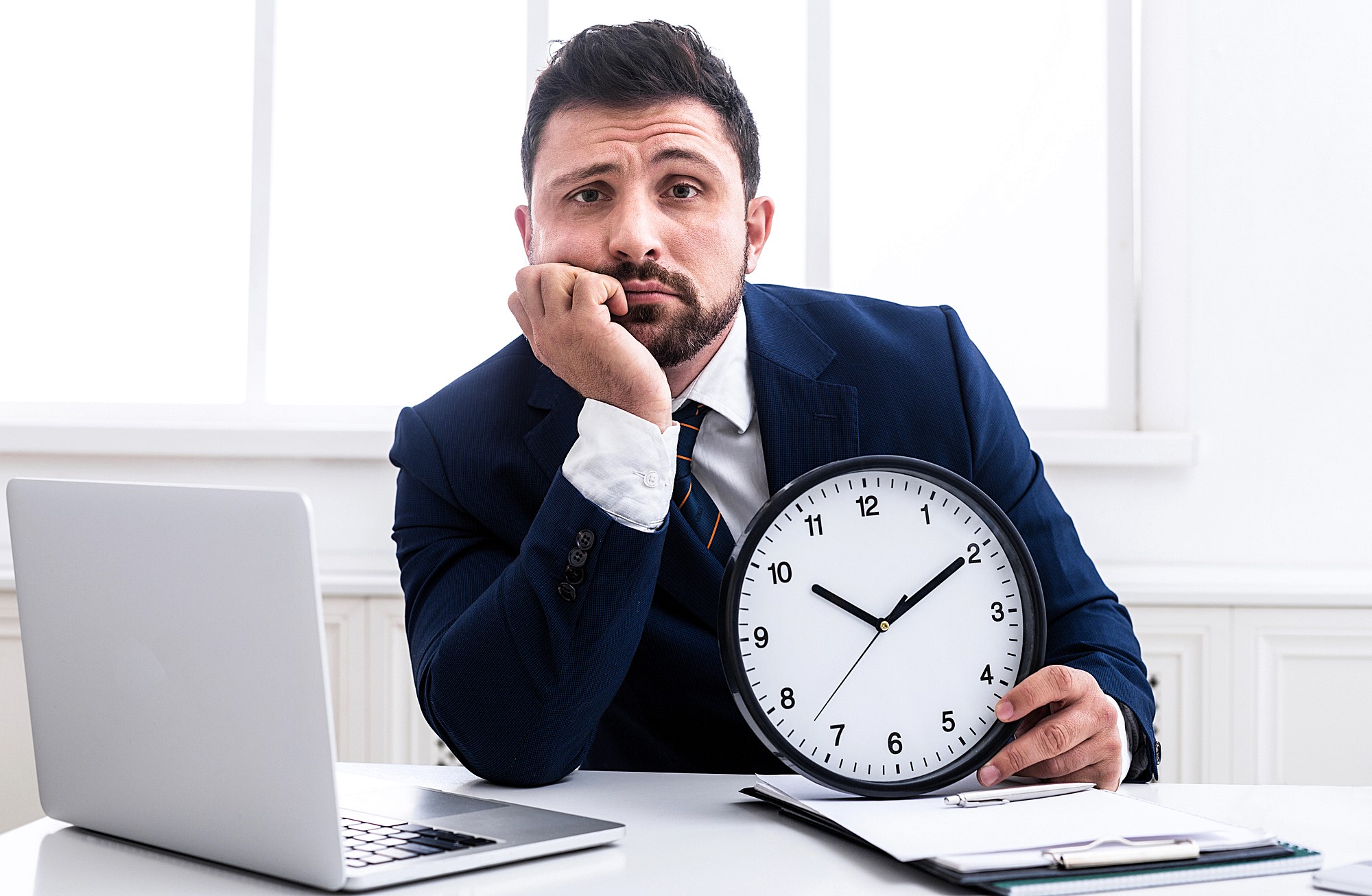 A man in a suit sits at a desk, resting his head on one hand and holding a wall clock with the other. A laptop and clipboard are in front of him. The expression on his face appears bored or impatient.