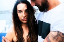 A woman with long brown hair stands closely with a man who has a beard and tattoos. She looks directly at the camera while the man wears a white t-shirt with a black square design. The background is blurred, suggesting an outdoor setting.