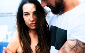 A woman with long brown hair stands closely with a man who has a beard and tattoos. She looks directly at the camera while the man wears a white t-shirt with a black square design. The background is blurred, suggesting an outdoor setting.