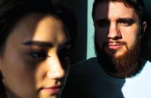 Close-up of a woman in the foreground and a man with a beard in the background, both partially illuminated by sunlight through blinds, casting dramatic shadows on their faces.