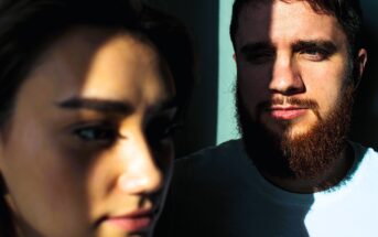 Close-up of a woman in the foreground and a man with a beard in the background, both partially illuminated by sunlight through blinds, casting dramatic shadows on their faces.