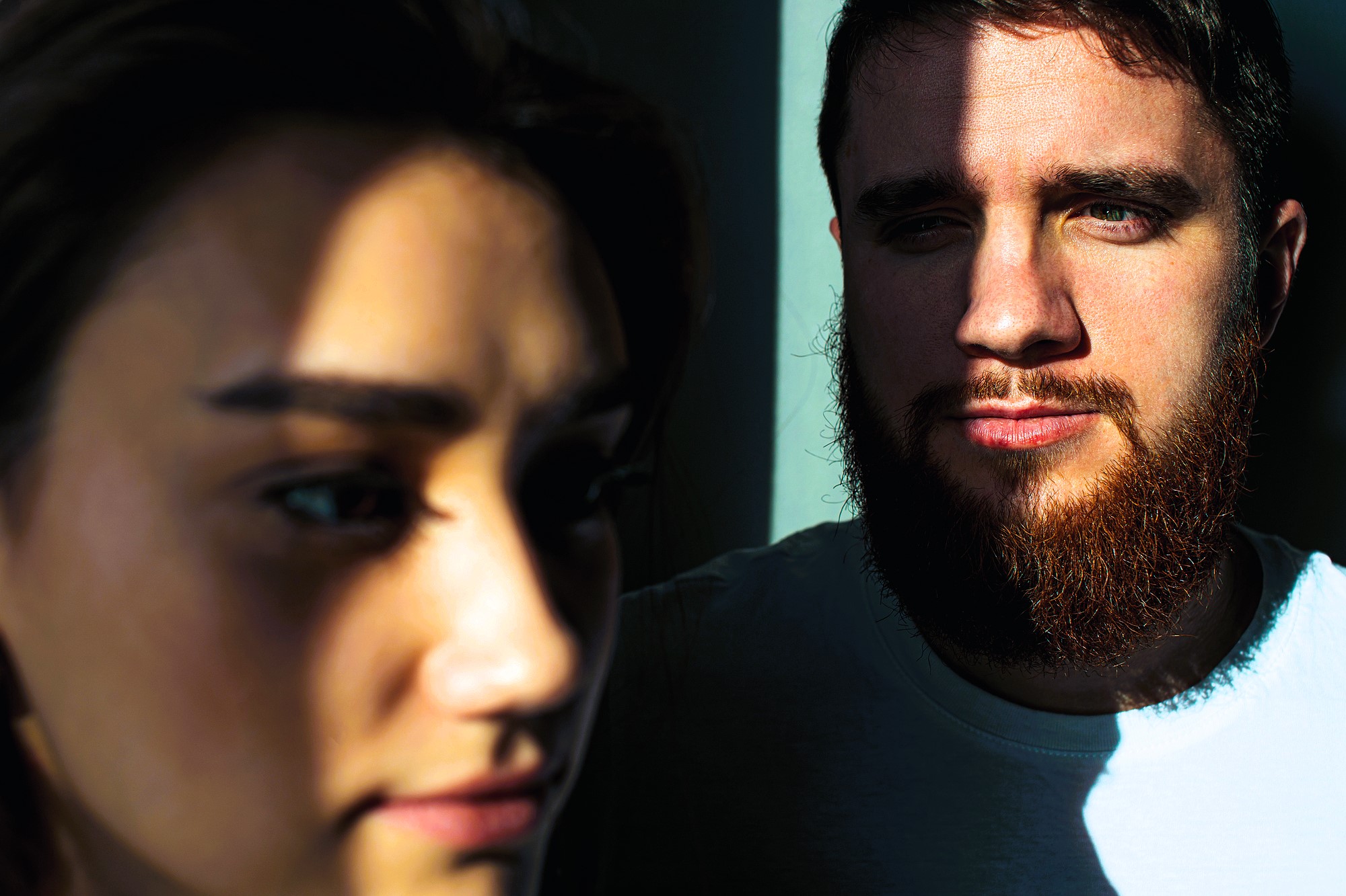 Close-up of a woman in the foreground and a man with a beard in the background, both partially illuminated by sunlight through blinds, casting dramatic shadows on their faces.