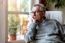 Elderly man wearing glasses and a gray sweater, resting his chin on his hand, gazing thoughtfully out a window. The room is softly lit with a potted plant visible in the background.