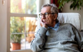 Elderly man wearing glasses and a gray sweater, resting his chin on his hand, gazing thoughtfully out a window. The room is softly lit with a potted plant visible in the background.