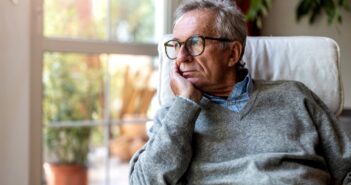 Elderly man wearing glasses and a gray sweater, resting his chin on his hand, gazing thoughtfully out a window. The room is softly lit with a potted plant visible in the background.