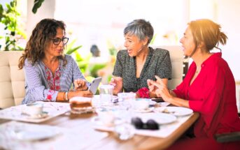 Three women sitting at an outdoor table, chatting and smiling. One woman shows something on her phone. The table has cups, saucers, and a partially eaten cake. They appear to be enjoying a pleasant conversation.