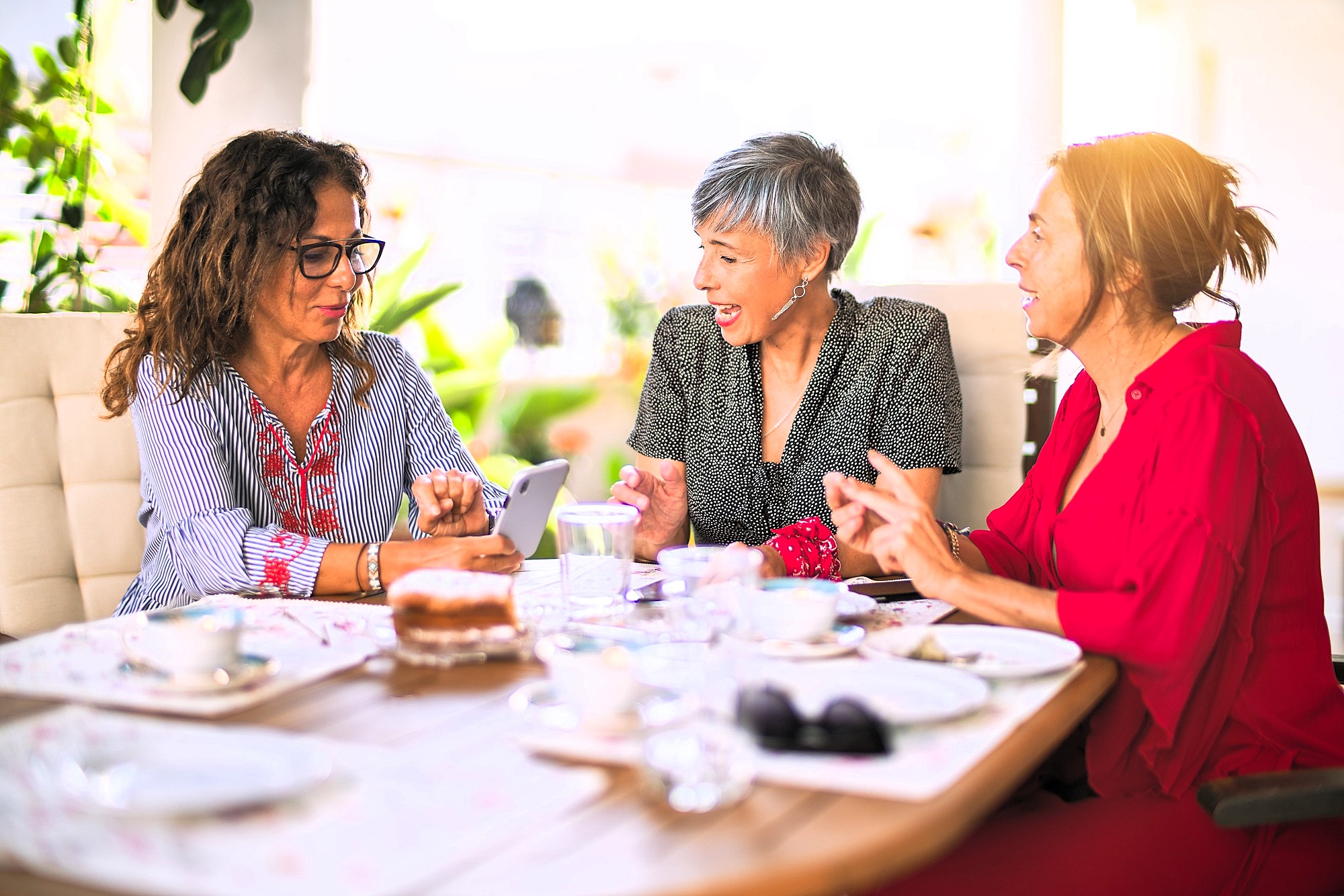 Three women sitting at an outdoor table, chatting and smiling. One woman shows something on her phone. The table has cups, saucers, and a partially eaten cake. They appear to be enjoying a pleasant conversation.