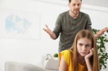 A man with a concerned expression gestures with his hands while a young girl in a yellow shirt sits in the foreground, looking away with a frustrated expression. They appear to be in a living room with a couch and plants.