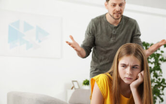 A man with a concerned expression gestures with his hands while a young girl in a yellow shirt sits in the foreground, looking away with a frustrated expression. They appear to be in a living room with a couch and plants.