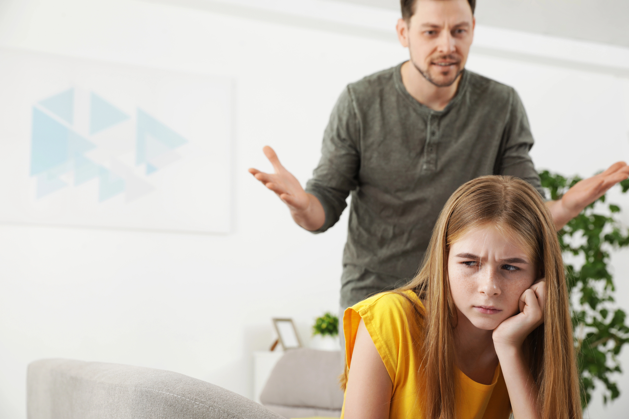 A man with a concerned expression gestures with his hands while a young girl in a yellow shirt sits in the foreground, looking away with a frustrated expression. They appear to be in a living room with a couch and plants.