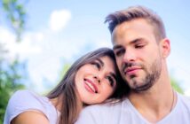 A young woman with long brown hair rests her head on a man's shoulder. Both are smiling slightly, wearing white shirts, and sitting outdoors against a blurred background of blue sky and greenery.